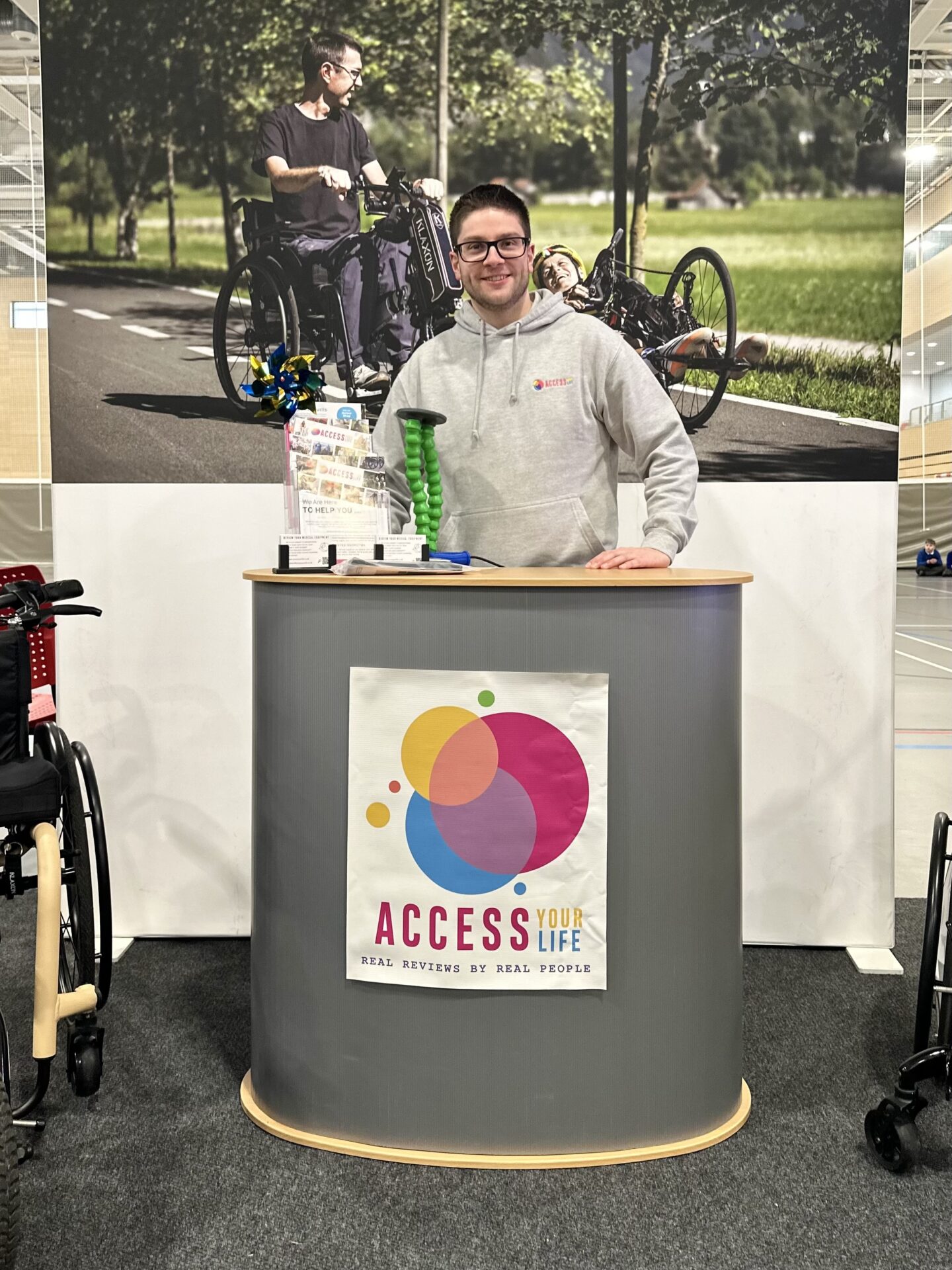 white man with dark hair and glasses stood behind a stand. the stand had a poster for access your life on. behind the man is an image of a man sat in a wheelchair with an adaption.