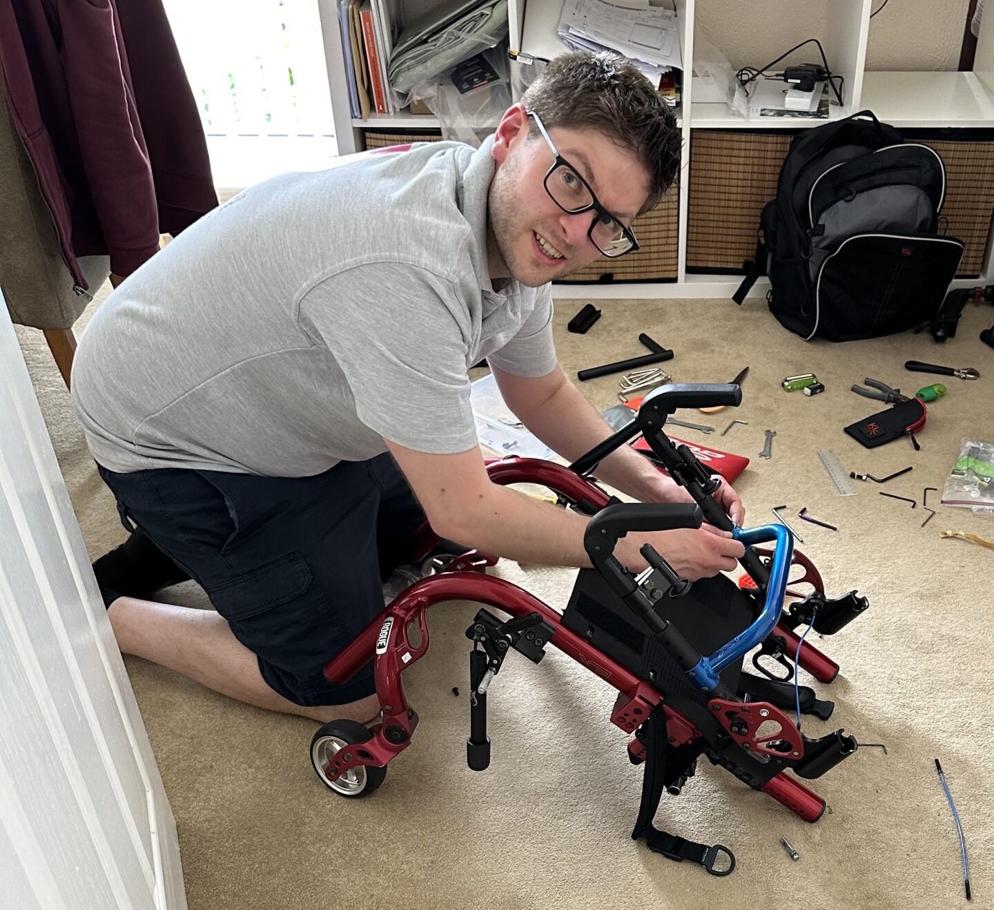 white man with dark hair and glasses knelt on the floor building a red wheelchair that has a blue handle on the back. in the back of the image you can see tools spread across the floor