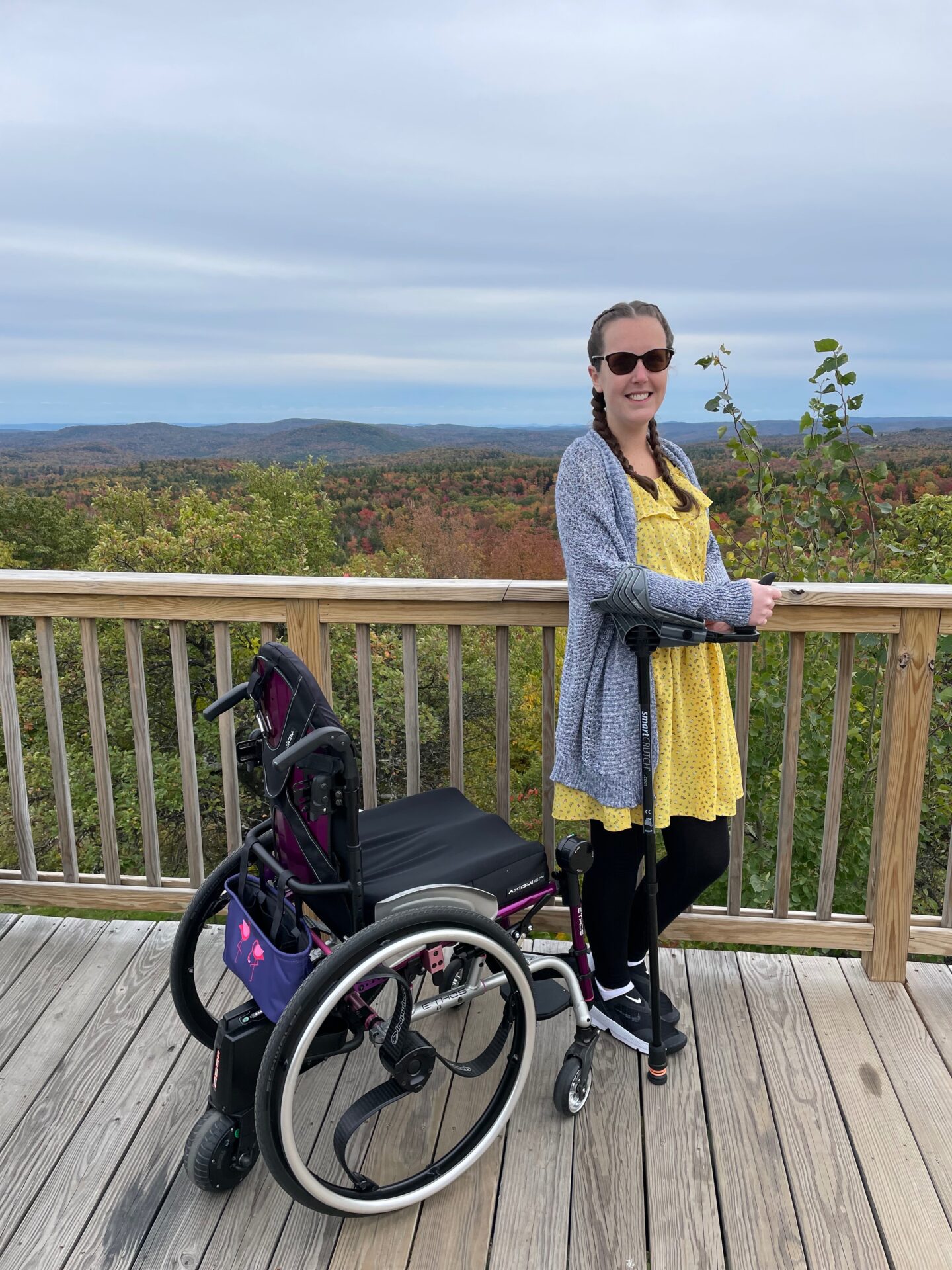 white woman with light brown hair wearing sunglasses dressed in a grey cardigan, yellow sun dress with black leggings and black trainers on using a crutch to support her. stood on a decking with a wooden railing her black and purple wheel chair are to the left of her.