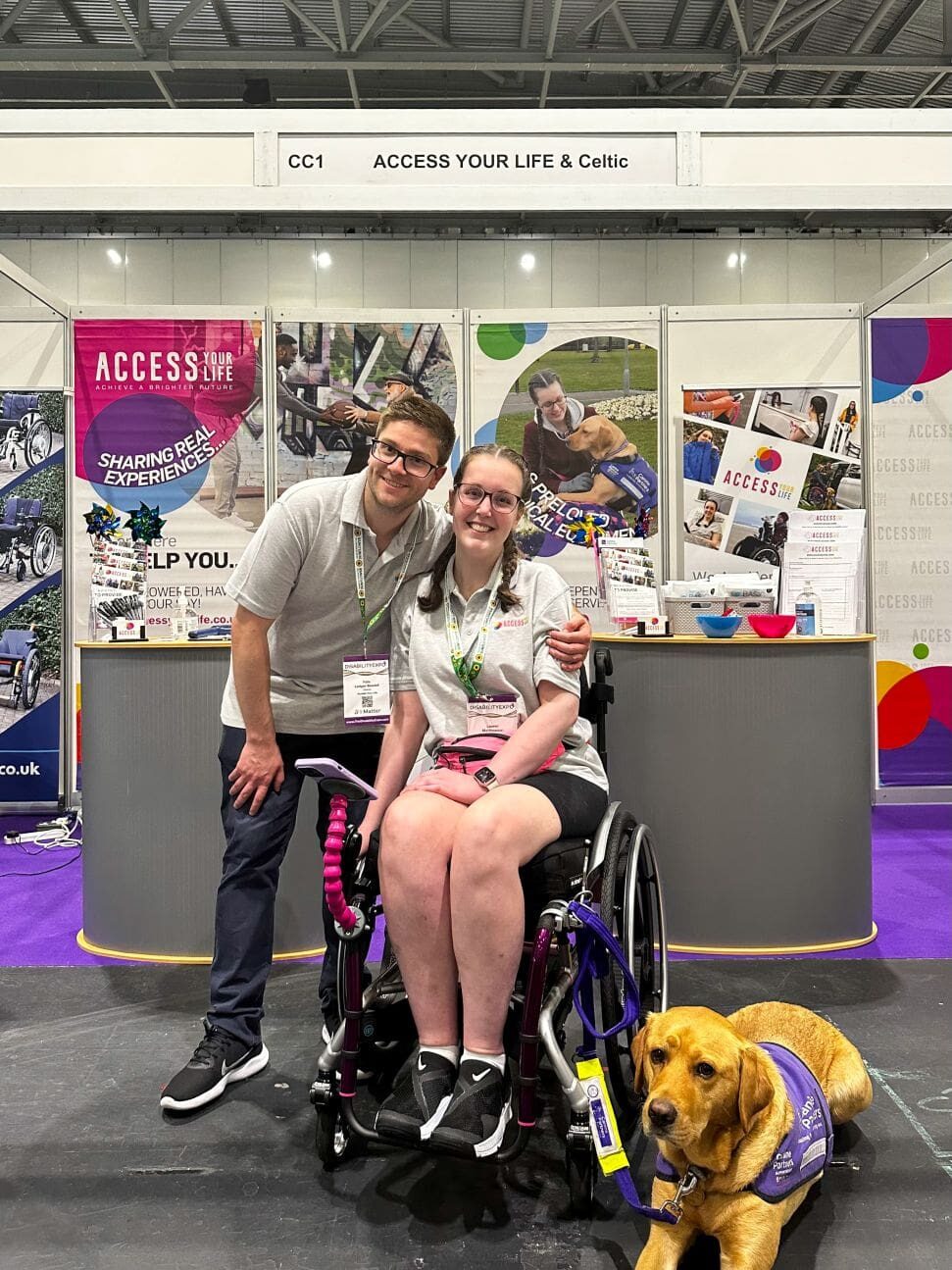 A man, woman in a wheelchair, & a service dog, posing for a photo in front of a display stand for 'Access Your Life'. From the lanyards they are wearing, we can infer that they are at some sort of convention, advertising their organisation.