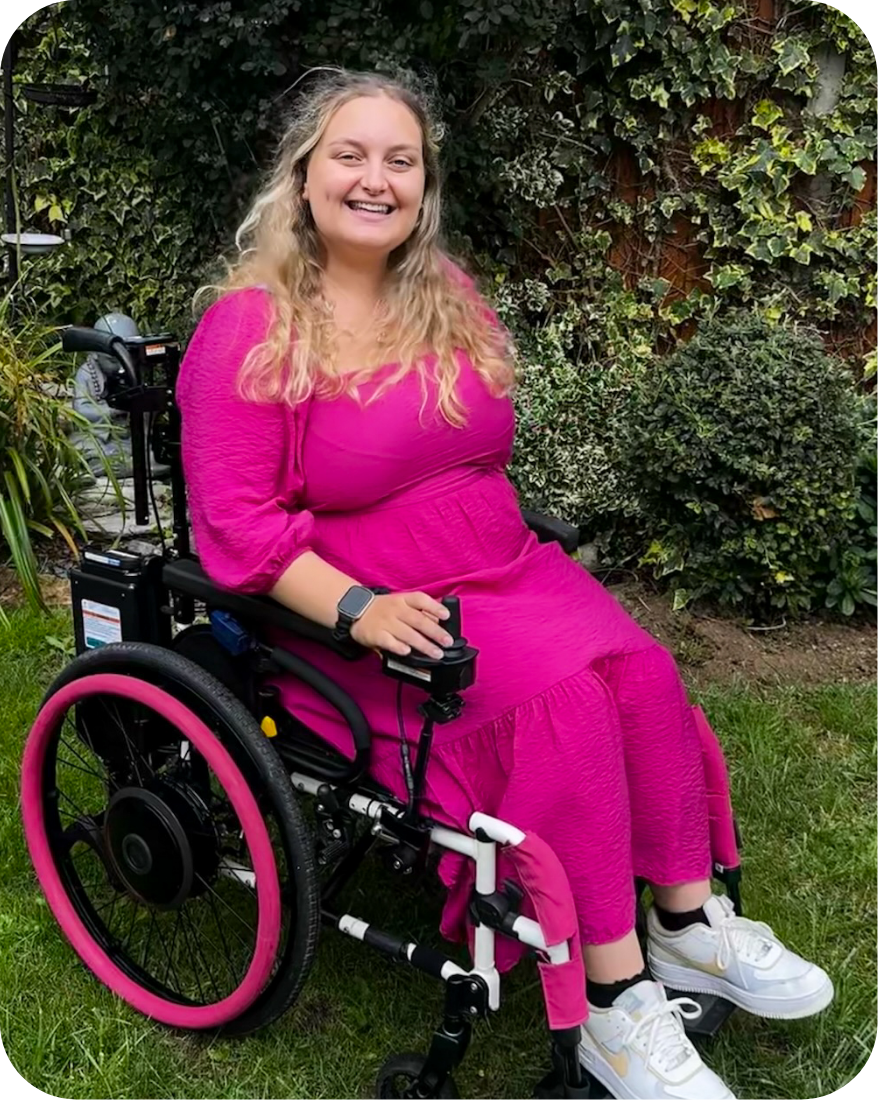 Photo of a smiling, blonde haired woman in an electric wheelchair. The woman is wearing a pink dress, pink sleeves, & matching pink colouring on her wheelchairs rims, with off colour white shoes. In the background, a garden & a statue of the Buddha can be seen.