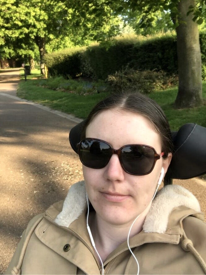 Selfie photo of a woman in a mobility aid, wearing sun glasses, wired ear buds, & a beige parka coat, during a day out at a park.