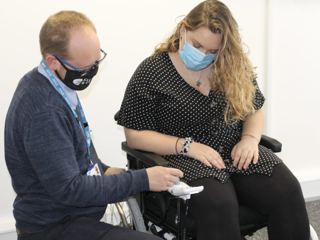 Picture of a blonde woman in an electric wheelchair, & a bespectacled man, with a receding blonde hairline, examining the joystick of the wheelchair. Both are wearing facemasks, & the man is wearing a NHS branded lanyard, from which we can infer he is some sort of medical professional.