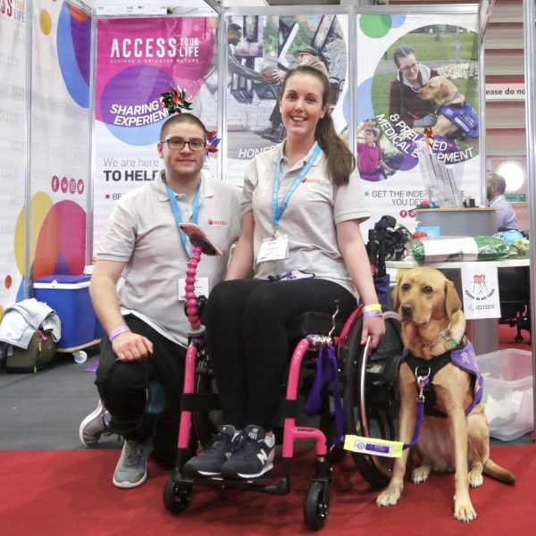 A man, a woman in a wheelchair, & a service dog, posing for a photo in front of a display stand for 'Access Your Life'. From the lanyards & matching shirts they are wearing, we can infer that they are at some sort of convention, advertising their organisation.