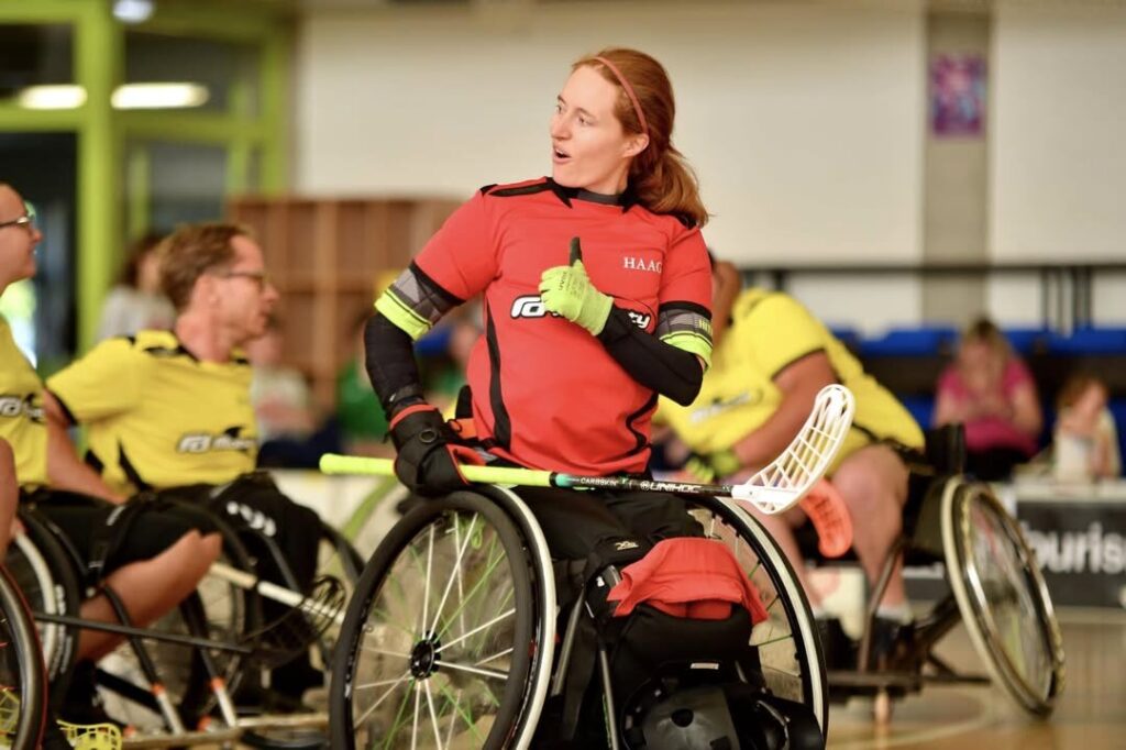 Photo focusing on a woman in a wheelchair due to having no lower legs, wearing a red jersey, & holding a hockey stick, as she is playing 'Wheelchair Hockey'. In the background other wheelchair hockey players in yellow shirts are seen.